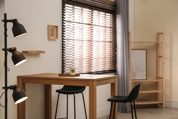 A modern home office with wooden Venetian blinds, a wooden desk, black chairs, and warm lighting, creating a stylish and functional workspace.