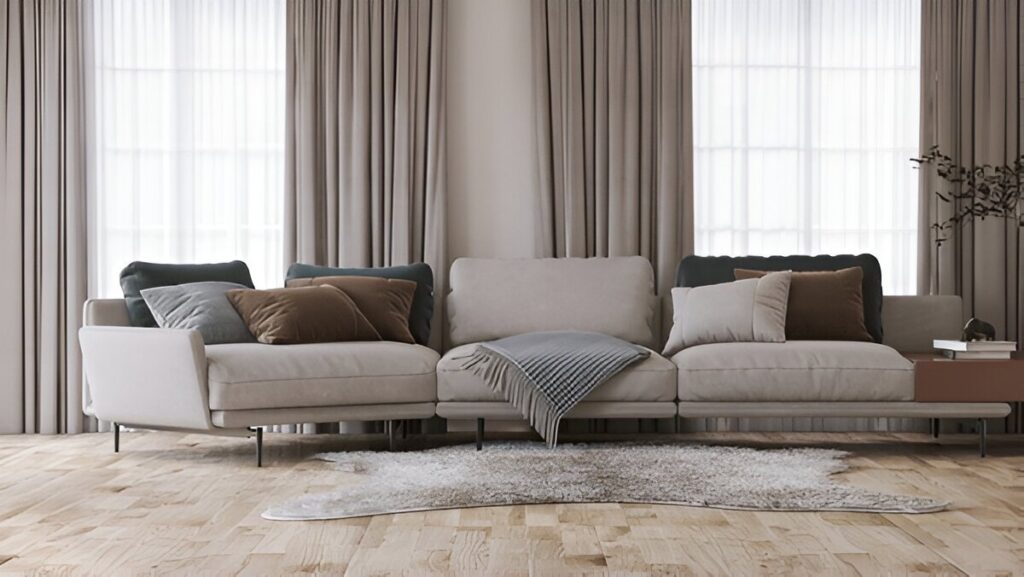 A modern living room with a beige sectional sofa, earthy-toned throw pillows, a gray throw blanket, a textured area rug, and white blackout curtains filtering natural light through large windows.