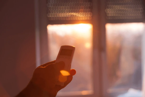 A hand holding a remote control to adjust motorized blinds in Dubai, with a beautiful sunset shining through the window.