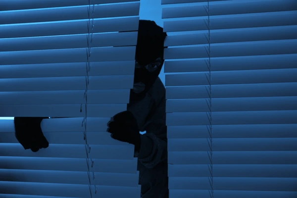 A masked figure peeking through broken blinds in a dark room, symbolizing scams and fraud in the Dubai window blinds market.