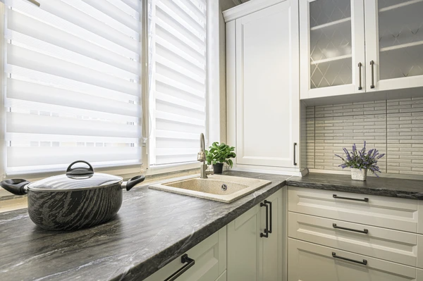 A modern kitchen featuring stylish Roman blinds 2025, offering privacy and elegance with a black marble countertop, white cabinets, and a clean aesthetic in Dubai.