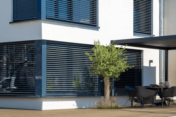 A contemporary home exterior featuring large windows with sleek black cut blinds, providing shade, privacy, and a modern aesthetic. The outdoor seating area and greenery enhance the stylish appeal.