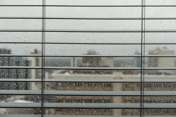 A rainy cityscape seen through horizontal window blinds, emphasizing the need for water-resistant blinds in humid and wet conditions in Dubai 2025.