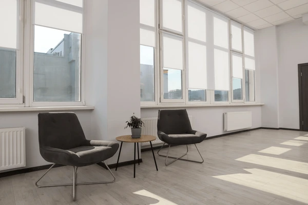 A bright, minimalist office space featuring large windows with sleek roller blinds, modern gray armchairs, and a stylish neutral color palette, showcasing how blinds offer superior light control and elegance over curtains.