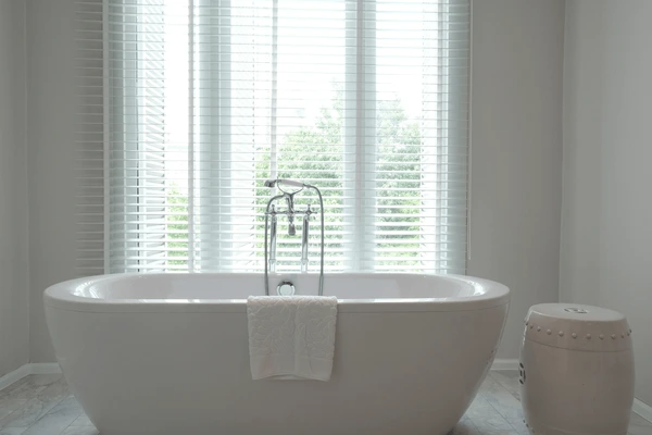 A minimalist bathroom featuring a freestanding bathtub with a sleek design, complemented by large windows covered with stylish white blinds for privacy.