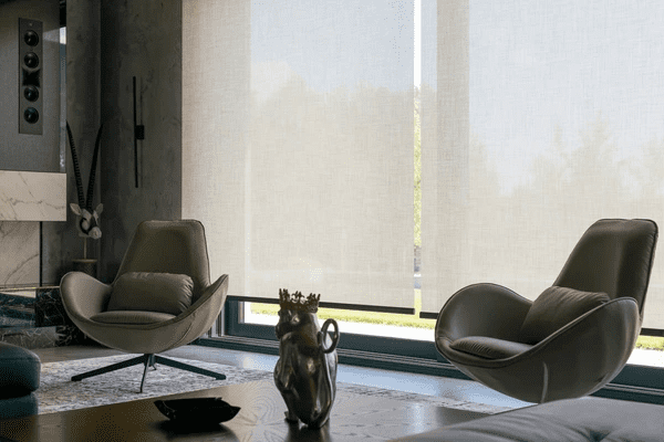 A stylish living room featuring beige soundproof roller blinds, two modern swivel chairs, and a marble fireplace, creating a peaceful and sun-protected interior.