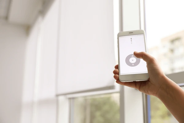A hand controlling motorized blinds in Dubai using a smartphone app, showcasing the convenience of smart home automation.