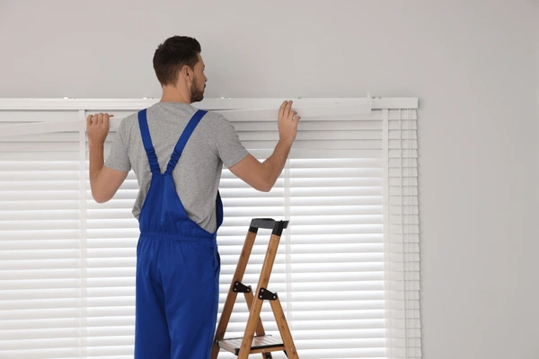 A professional worker in a blue uniform installing white horizontal blinds using a ladder, ensuring a precise and secure fit for a modern home in Dubai.