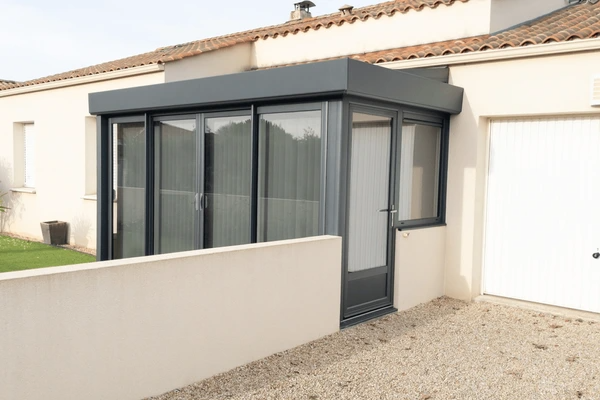 A small modern conservatory attached to a house, featuring grey frames and vertical blinds inside the glass panels.