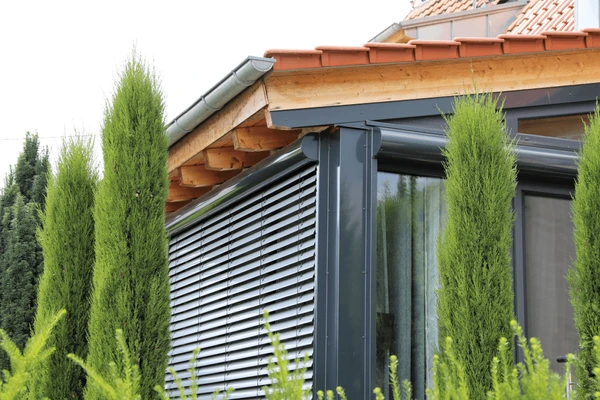 Conservatory with dark-colored exterior Venetian blinds surrounded by lush green trees and a tiled roof.