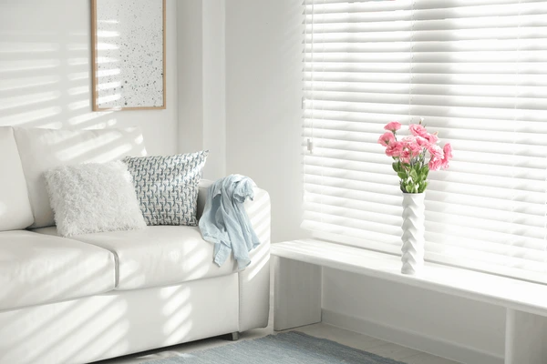 Bright living room in Dubai with white horizontal blinds, cozy sofa, and a white vase of pink flowers creating a luxurious and warm ambiance in 2025.
