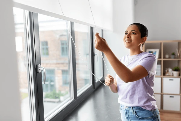 Smiling woman adjusting sleek white cord blinds in a bright, modern living space with large windows.