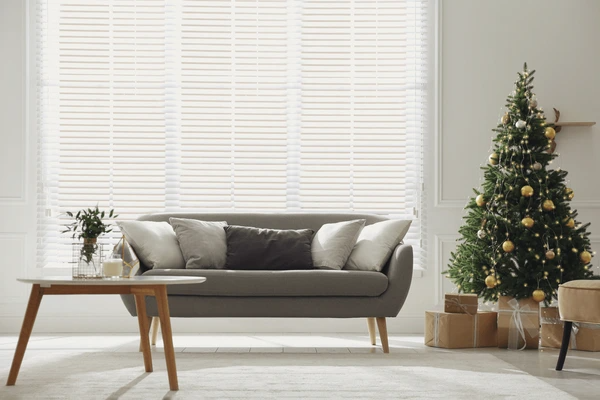 Elegant living room in Dubai with white blinds, grey sofa, and decorated Christmas tree beside wrapped gifts.