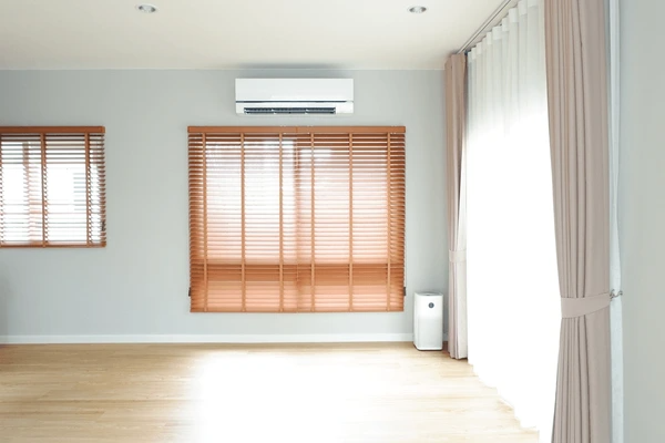 Spacious living room with long wooden blinds on two windows, soft beige curtains, and minimalist décor including a wall-mounted air conditioner.