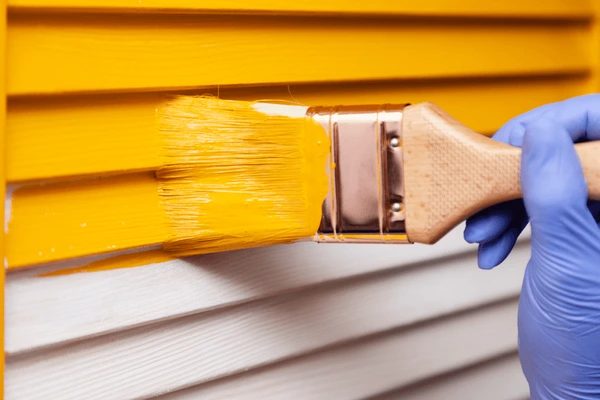 Close-up of a gloved hand painting window blinds yellow, showcasing the best painted blinds trend in Dubai 2025 for stylish interiors.