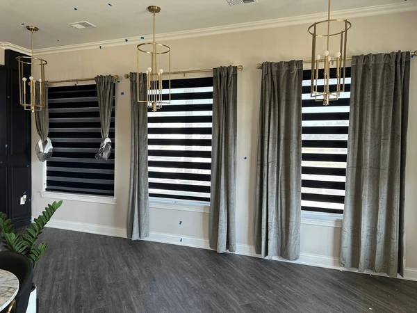 Elegant living room with black zebra blinds and grey velvet curtains on large windows, under gold pendant lights.