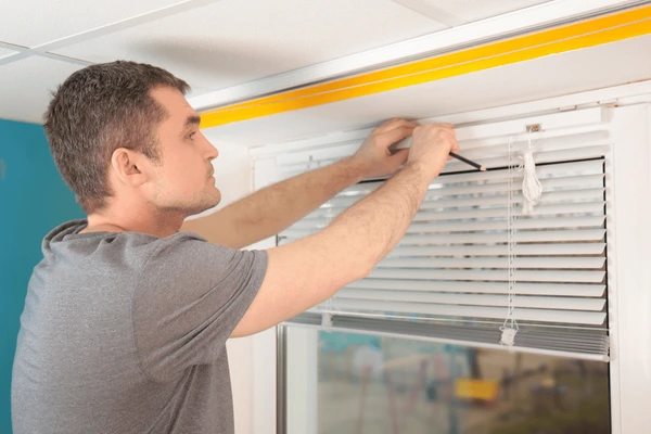 Man repairing white venetian blinds using a screwdriver on a bright window frame.