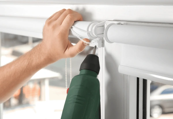 Close-up of a hand repairing white roller blinds with a power drill in a modern Dubai home.