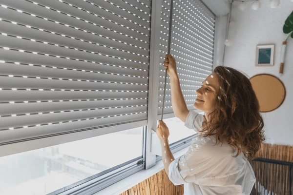 Smiling woman closing modern roller shutters in a bright indoor balcony, showcasing sleek and trendy window coverings perfect for Dubai homes in 2025.