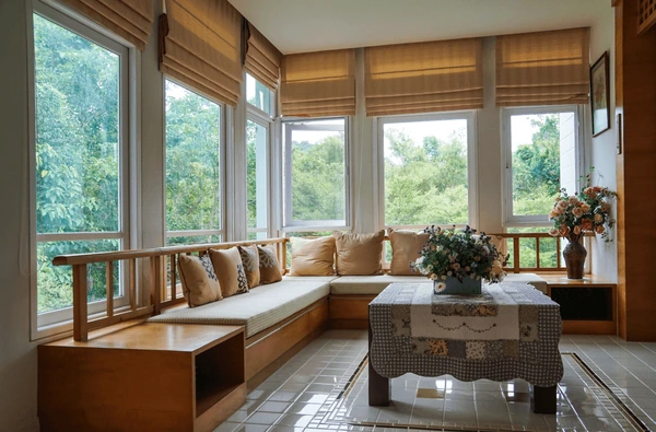 Cozy living room with floor-to-ceiling windows dressed in brown Roman blinds, surrounded by wooden furniture and lush outdoor views.