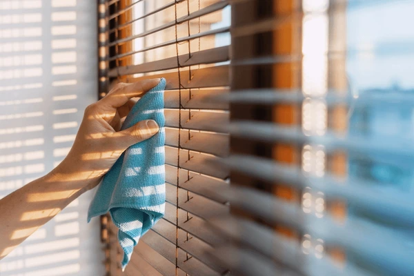 Hand holding a blue cloth, cleaning wooden blinds in warm sunlight, showing the importance of blind maintenance for Dubai homes.