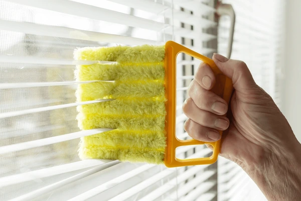 A close-up of a hand using a yellow microfiber blind cleaner to wash horizontal window blinds in natural daylight.