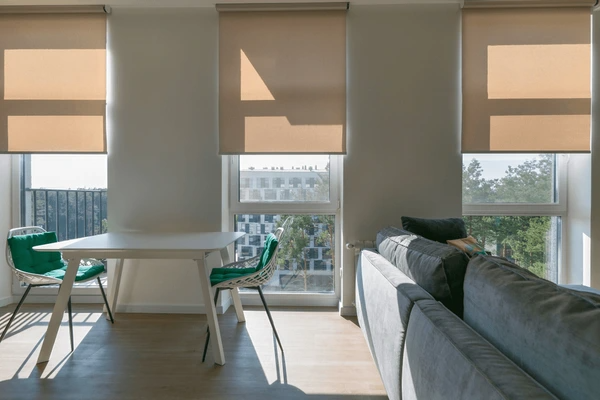 Sunlit living and dining area with beige motorized roller blinds, green accent chairs, and a modern grey sofa in a Dubai apartment, 2025.
