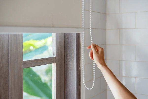 A hand pulling the chain of a sleek roller blind in a bright, tiled interior, offering smooth light control and privacy.