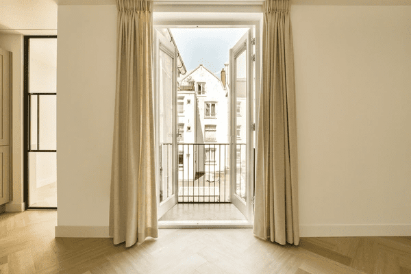 Sunlit room with beige thermal curtains framing open balcony doors, revealing traditional buildings outside.