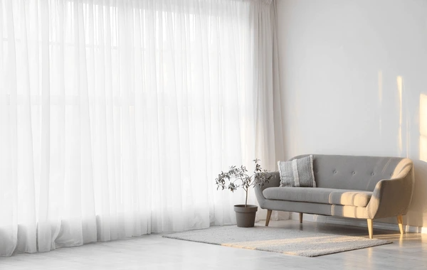 Best Drop Curtains in Dubai 2025 - Light, airy curtains complementing a minimalist living room with a gray sofa, potted plant, and soft natural lighting.