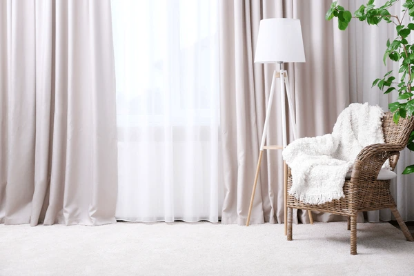 Best Drop Curtains in Dubai 2025 - Soft, luxurious beige drop curtains complementing a cozy chair, floor lamp, and green plant in a modern living room.