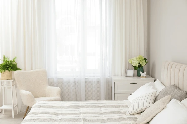 Soft white noise-reducing curtains installed in a modern cozy bedroom with a neutral-toned bed and accent chair in Dubai.