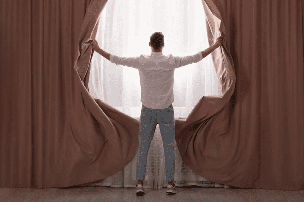 Man opening elegant dusty rose curtains to reveal sheer white drapes in a bright, modern room.