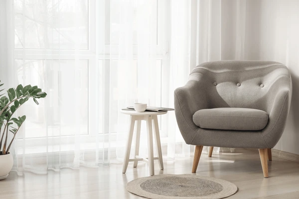 Modern cozy living room with sleek gray armchair, sheer white curtains, round rug, and indoor plant – showcasing top-rated sheer curtain trends for Dubai homes in 2025