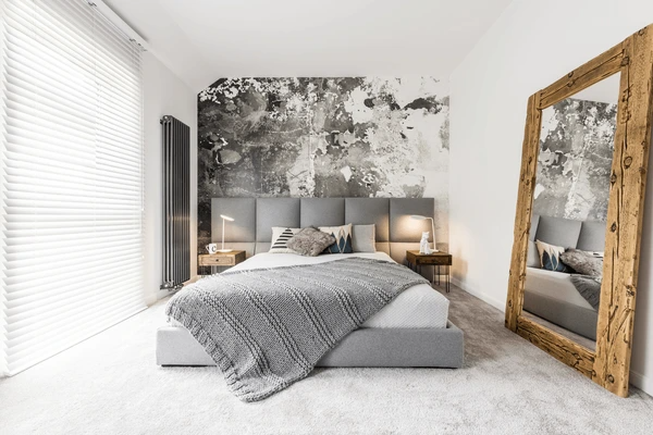 Modern bedroom with grey upholstered bed, textured accent wall, large wooden mirror, and sleek white stylish blinds filtering natural light.
