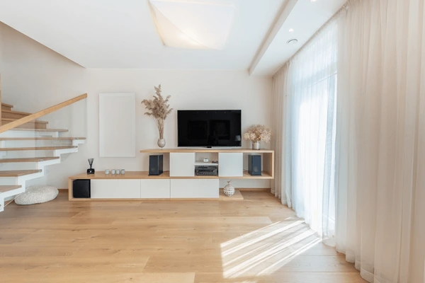 Bright modern living room with beige unwrinkled curtains, a floating staircase, wooden floors, and sleek entertainment setup.