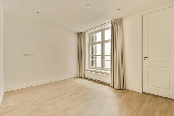Bright, empty room with beige unwrinkled curtains, white walls, wood flooring, and a large window letting in natural light.