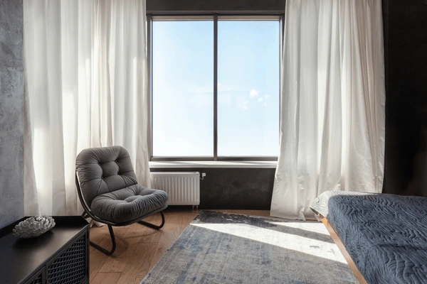 Modern cozy bedroom featuring soft unwrinkled curtains, a plush grey lounge chair, and a minimalist decor with sleek dark accents.