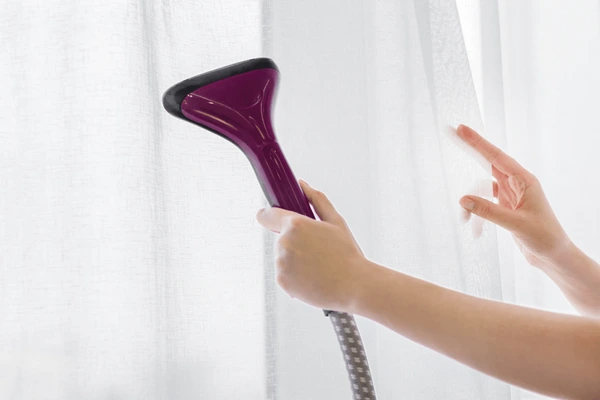 Close-up of hands steaming white curtains with a handheld garment steamer for a smooth, wrinkle-free finish.