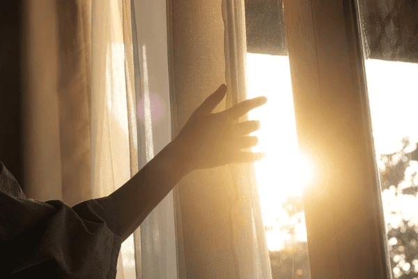 A hand opens soft, beige thermal curtains in warm morning sunlight, capturing the cozy elegance of heat-retaining drapes for modern Dubai interiors.