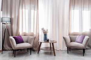Elegant living room with luxurious taupe and white curtains, modern armchairs with purple pillows, a wooden table, and a floor lamp.