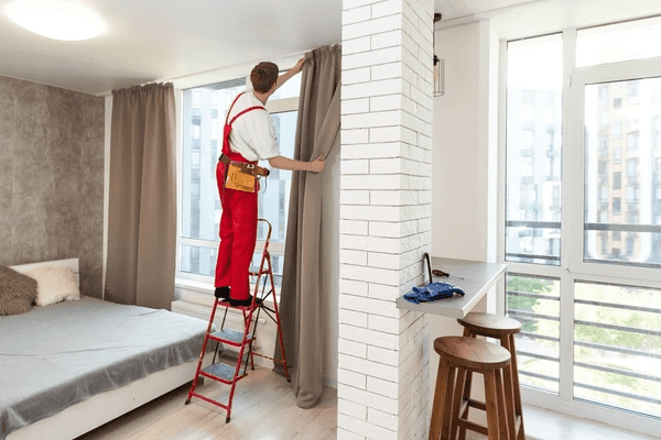 Professional technician installing modern grey curtains in a stylish Dubai apartment bedroom with floor-to-ceiling windows.
