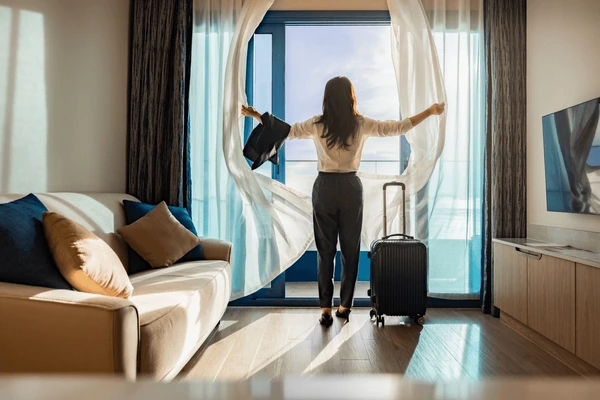Woman opening sheer white curtains in a modern Dubai apartment with sunlight streaming in through a balcony window.