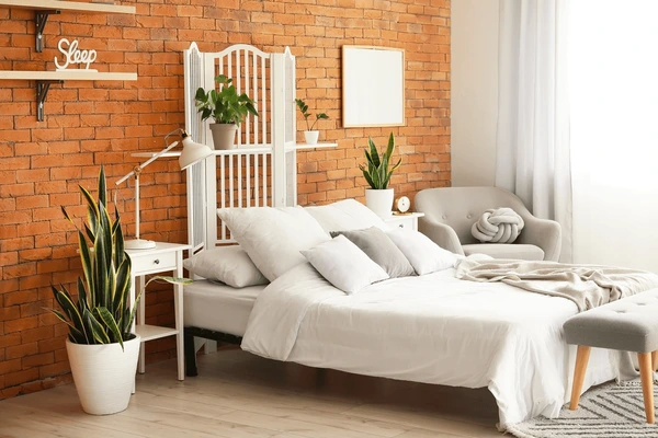 Stylish bedroom with exposed brick wall, white bed, indoor plants, and affordable white curtains filtering sunlight.