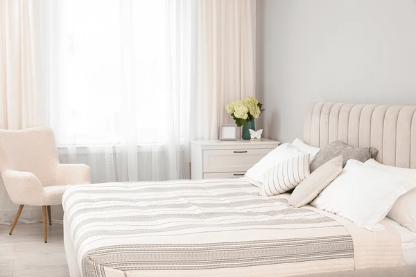 Bright bedroom with large cream-colored curtains behind a stylish upholstered bed, soft beige tones, and natural light.