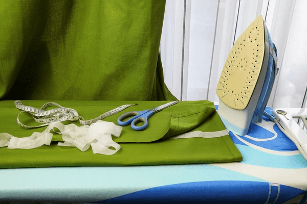 Professional curtain-making setup with green fabric, scissors, measuring tape, and an iron, symbolizing expert craftsmanship in Dubai.

