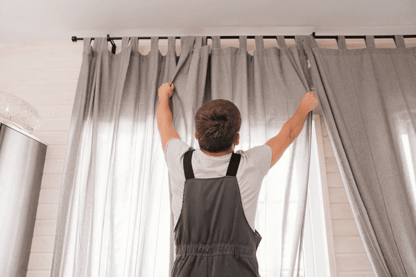 A professional handyman in uniform installing gray curtains on a black curtain rod in a bright modern room in Dubai 2025.

