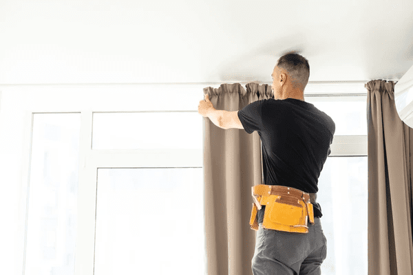 Professional handyman installing beige curtains in a modern Dubai apartment, showcasing Emirati Blinds & Curtains expertise.