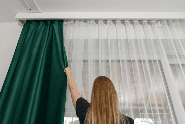A woman closing thick green blackout curtains layered over white sheer curtains in a modern Dubai home interior.

