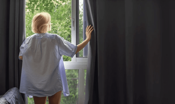 Woman opening blackout curtains in a cozy Dubai bedroom, revealing lush greenery outside while maintaining modern indoor privacy and stylish comfort.

